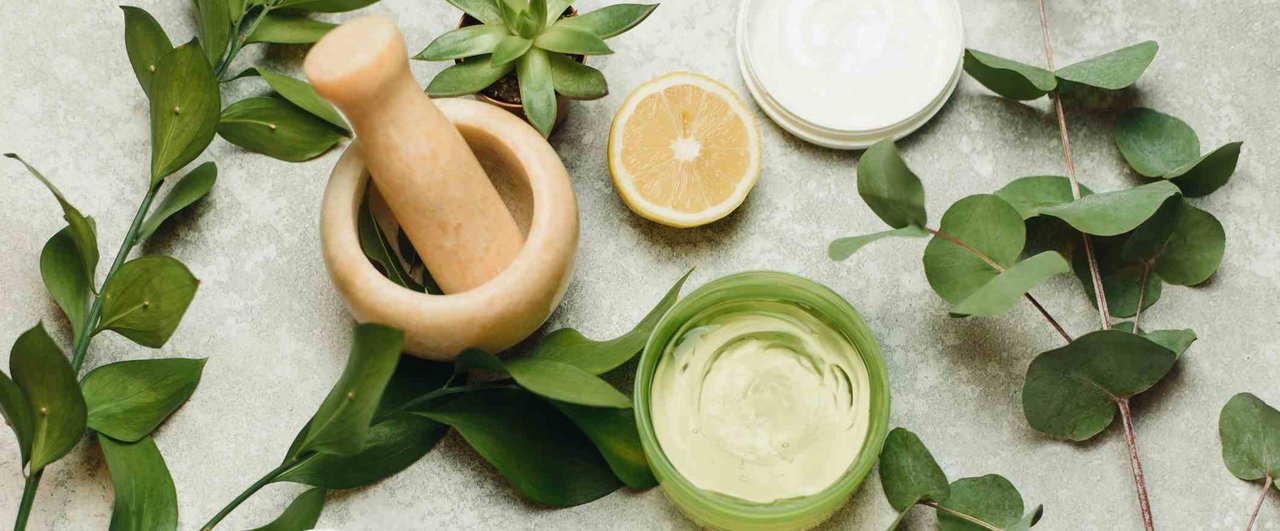 Natural emollience and sustainable beauty ingredients, including a mortar, leaves, lemon, and cream in a green jar.