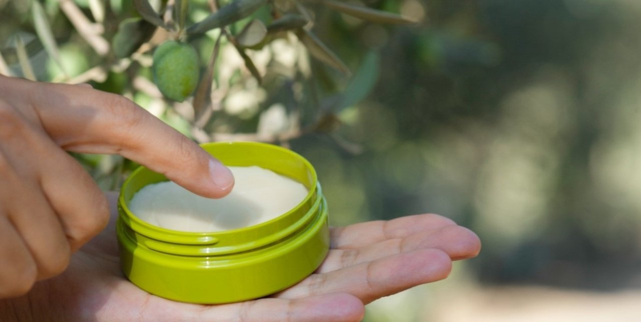 A hand holds an open green jar of cream with a finger dipping in, against an olive tree, symbolizing emollience and natural formulations.