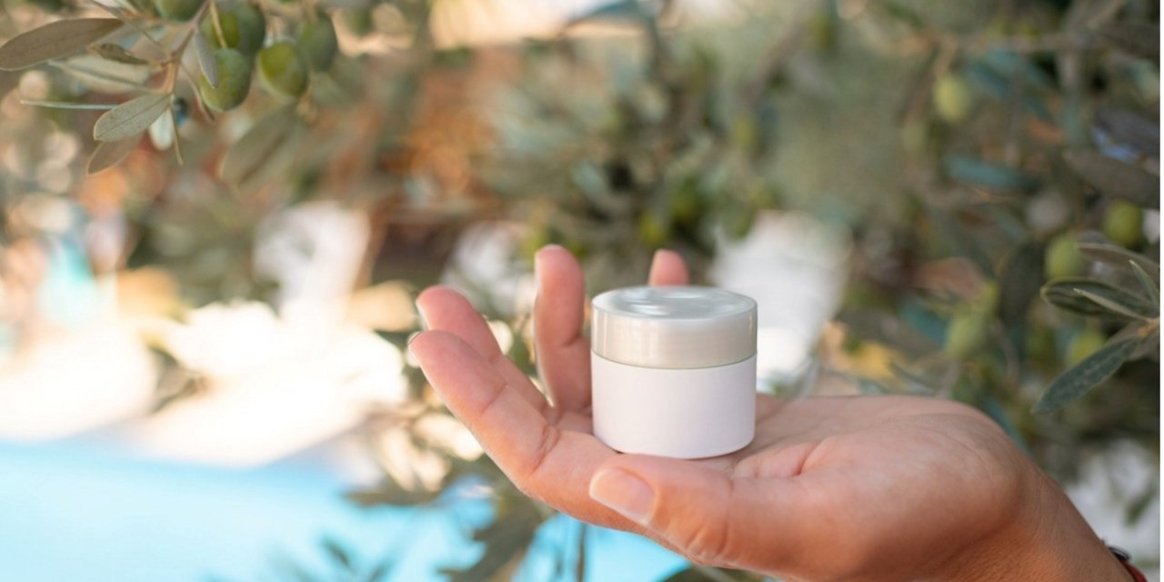 A hand holds a white cosmetic jar with an olive tree in the background, symbolizing natural formulations and beauty ingredients.